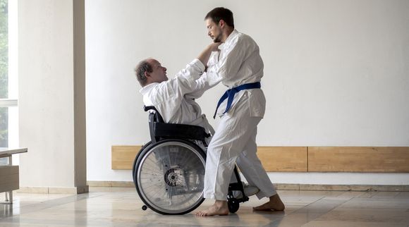 Zwei Männer in weißen Karateanzügen trainieren miteinander. Einer ist Rollstuhlfahrer, der andere nicht.